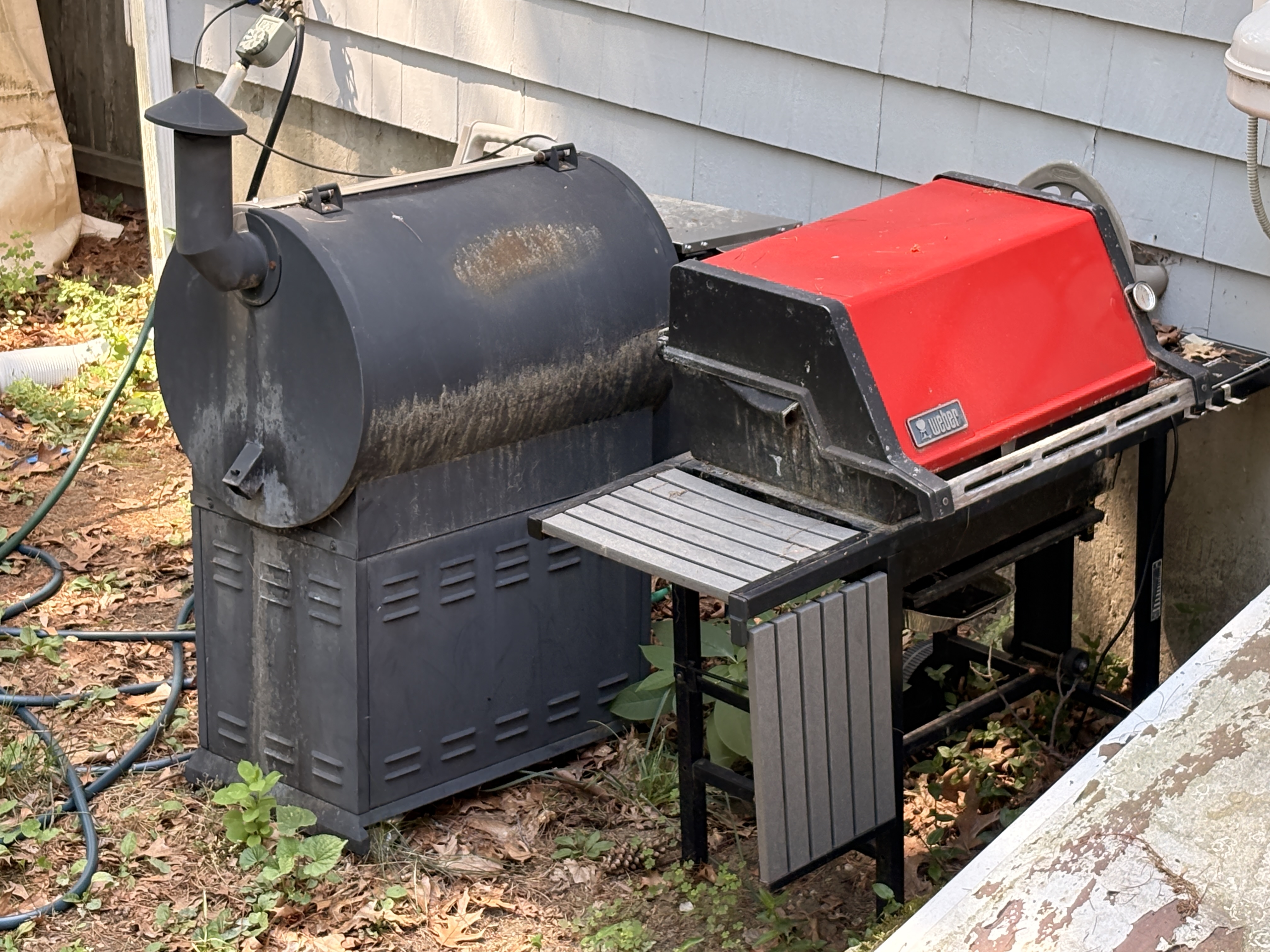 Older red and black Weber gas grill, alongside the barrel smoker